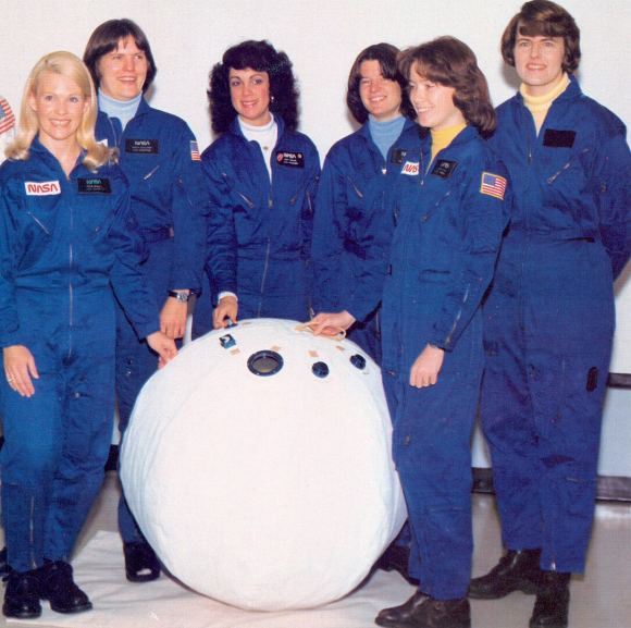 Шість жінок-космонавтів у синіх скафандрах стоять навколо білої рятувальної кулі.